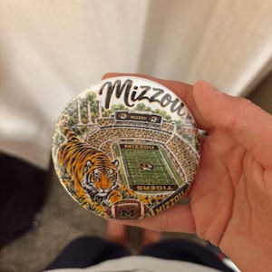 May include: A round pinback button featuring a graphic illustration of a football stadium with the text Mizzou at the top. A tiger is positioned in the foreground, and a football with the letter M is at the bottom. The stadium field displays the text Mizzou and Tigers.
