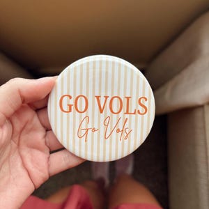 Puede incluir: Una chapa redonda con un patrón de rayas verticales en color crema y blanco. El texto GO VOLS está impreso en letras mayúsculas de color naranja, con la frase Go Vols escrita debajo en una fuente naranja más pequeña y cursiva.