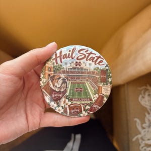 May include: A round button featuring a graphic of a bulldog, a football stadium, and a football. The text "Hail State" is printed in maroon at the top, with "BULLDOGS" at the bottom. The design includes maroon and white floral accents.