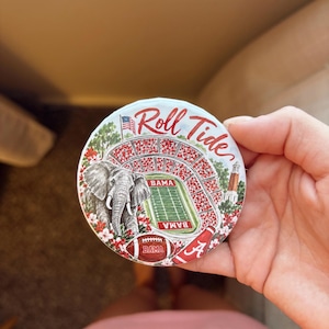 May include: A round ceramic coaster featuring a colorful illustration of a football stadium, a grey elephant, a football, and a megaphone. The text Roll Tide is written in red script at the top, with BAMA printed on the field and football.