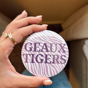 May include: A circular button featuring a purple and white zebra stripe pattern. The text GEAUX TIGERS is printed in bold, dark purple capital letters across the center of the design. The button has a smooth, glossy finish and a round shape.