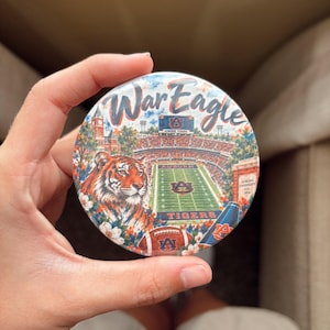 May include: A circular button featuring a colorful illustration of a football stadium, a tiger, and a football. The text War Eagle is printed at the top in a script font. The design includes orange and blue colors, a brick building, and flowers.