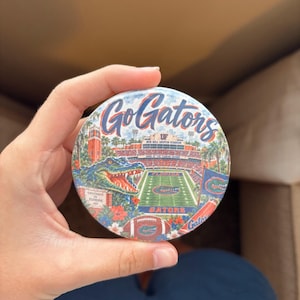 May include: A circular pinback button featuring the text Go Gators in blue script. The graphic depicts Ben Hill Griffin Stadium, a large alligator head, a football field with the Florida logo, a football, and tropical flowers in orange and blue.