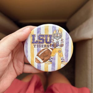 May include: A round button featuring a watercolor-style background of vertical purple and gold stripes. The design includes the text LSU TIGERS in purple, a brown football, and a cartoon tiger mascot wearing a football uniform and helmet.