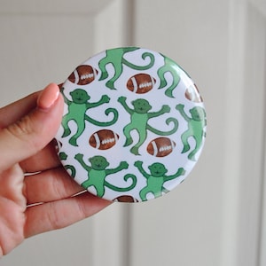 May include: A circular button featuring a repeating pattern of light green cartoon monkeys and brown American footballs on a white background. The design is printed on a smooth, glossy surface with a metallic edge, showcasing a playful motif.
