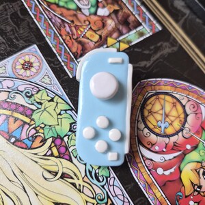 May include: A light blue controller-shaped charm with white buttons and a joystick. The charm is set against a backdrop of colorful stained-glass artwork featuring a video game character.
