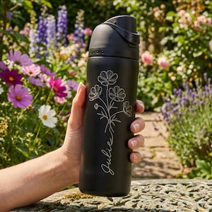 May include: Black insulated water bottle with a floral design and the name "Julie" in script. The bottle has a black cap with a handle and the brand name "awala" printed on the side. The background features a garden with colorful flowers.
