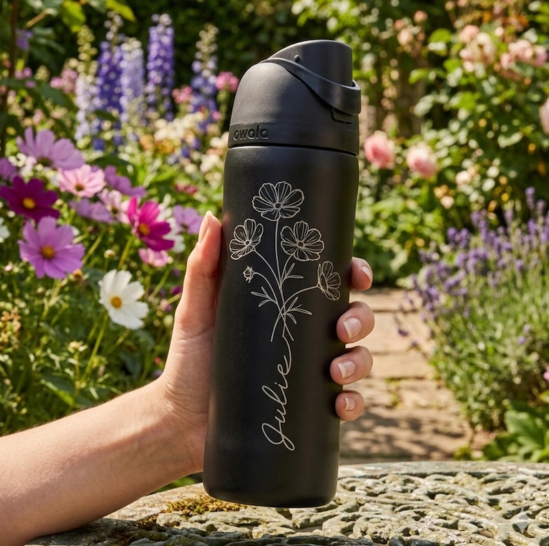 May include: Black insulated water bottle with a floral design and the name "Julie" in script. The bottle has a black cap with a handle and the brand name "awala" printed on the side. The background features a garden with colorful flowers.