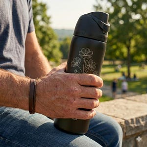 May include: A black water bottle with a floral design. The bottle has a flip-top lid and is held by a person wearing a brown leather bracelet and blue jeans. The bottle has the word "owala" printed on it.