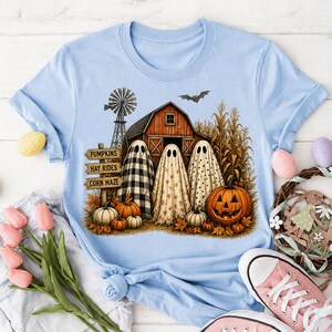 Può includere: Una t-shirt azzurra con una grafica raffigurante un fienile rosso, un mulino a vento, steli di mais e tre fantasmi drappeggiati in lenzuola a motivi. Un cartello in legno riporta le scritte PUMPKINS, HAY RIDES e CORN MAZE. Una zucca è in primo piano.