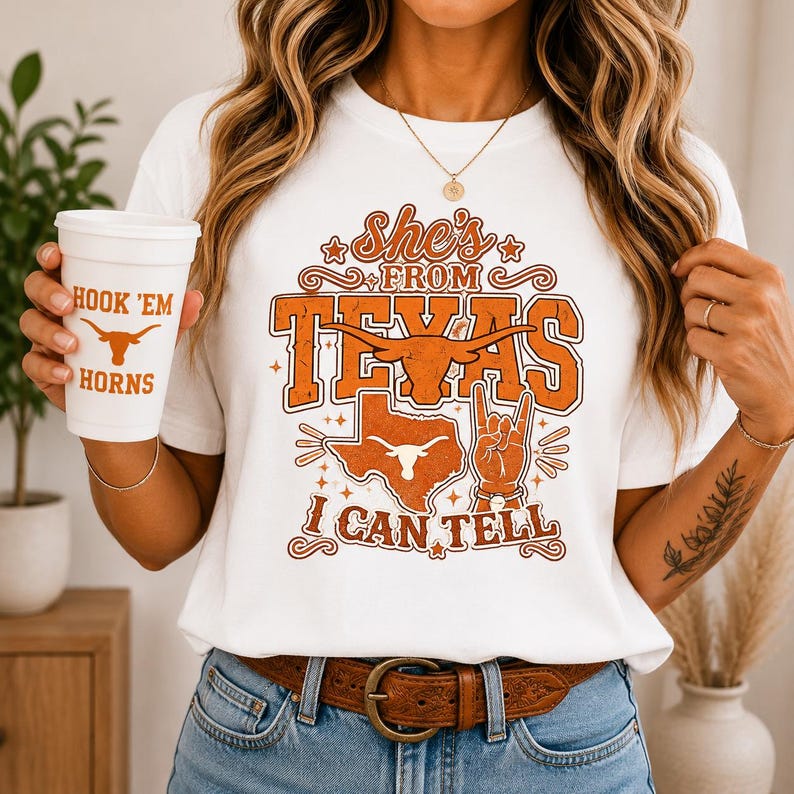 May include: A white t-shirt featuring orange graphic text that reads She's FROM TEXAS I CAN TELL, a longhorn steer silhouette, a map of Texas, and a hand gesture. A white cup with the orange text HOOK 'EM HORNS and a longhorn steer icon.