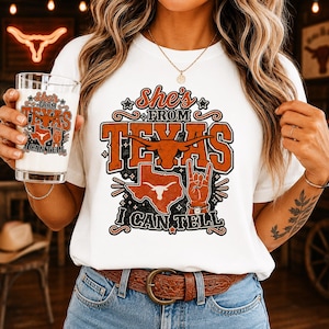 May include: A white t-shirt featuring a graphic design with the text "She's from Texas I can tell" in orange and black. The design includes a longhorn skull, a map of Texas, and a hand making a rock-and-roll gesture. A glass with the same design.