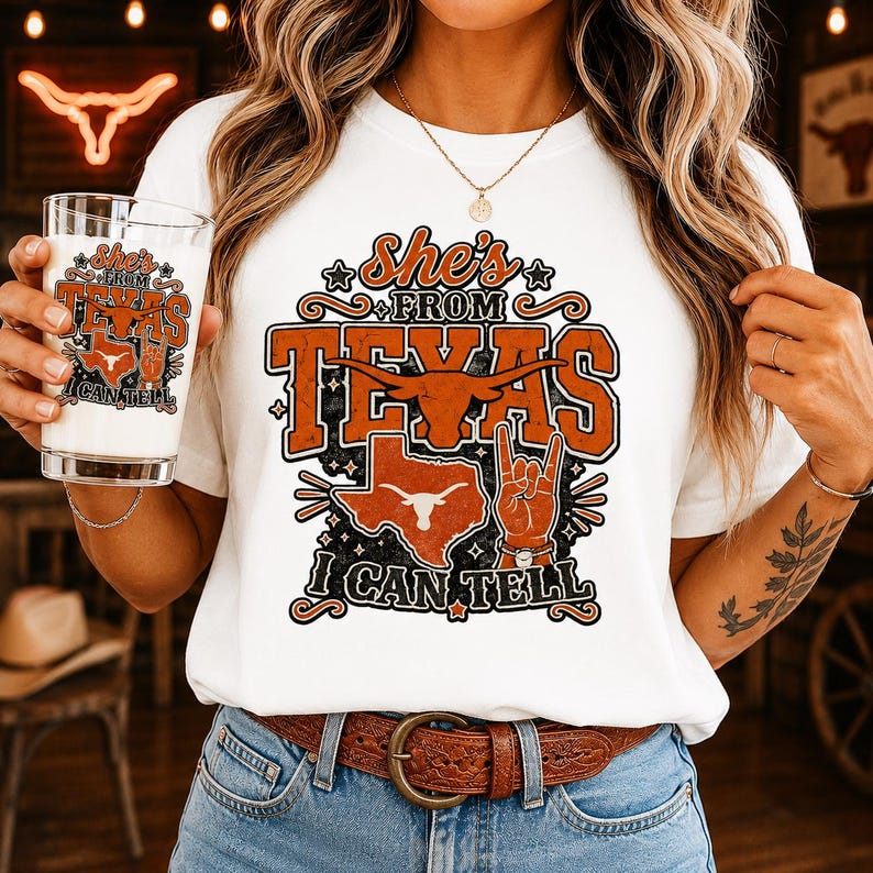 May include: A white t-shirt featuring a graphic design with the text "She's from Texas I can tell" in orange and black. The design includes a longhorn skull, a map of Texas, and a hand making a rock-and-roll gesture. A glass with the same design.