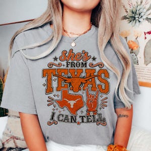 May include: A grey short-sleeve t-shirt featuring a graphic print in orange and black. The text reads She's FROM TEXAS I CAN TELL. The design includes a longhorn steer head, a map of Texas, and a hand making a hook 'em horns gesture.