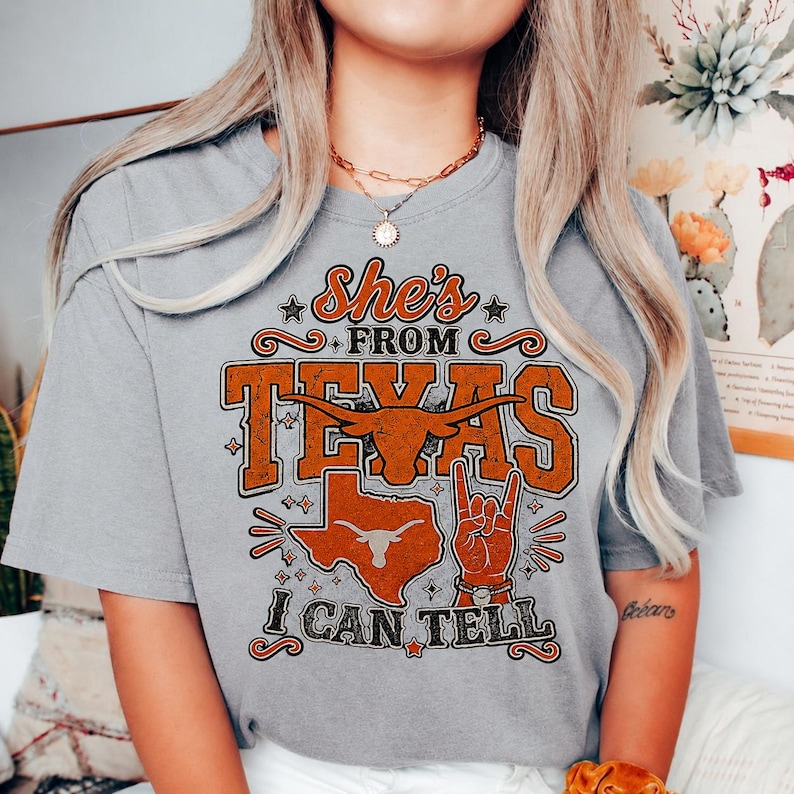May include: A grey short-sleeve t-shirt featuring a graphic print in orange and black. The text reads She's FROM TEXAS I CAN TELL. The design includes a longhorn steer head, a map of Texas, and a hand making a hook 'em horns gesture.
