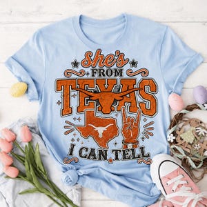May include: A light blue t-shirt featuring the orange text "She's FROM TEXAS I CAN TELL" with a longhorn steer graphic, a map of Texas, and a hand making a "hook 'em horns" gesture. The design includes decorative stars and flourishes.