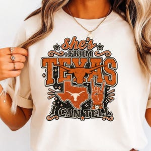 May include: A cream-colored t-shirt featuring a graphic print with the text "She's FROM TEXAS I CAN TELL". The design includes a burnt orange longhorn silhouette, a map of Texas, and a hand making a "hook 'em" gesture, all in orange and black.