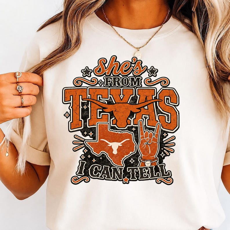 May include: A cream-colored t-shirt featuring a graphic print with the text "She's FROM TEXAS I CAN TELL". The design includes a burnt orange longhorn silhouette, a map of Texas, and a hand making a "hook 'em" gesture, all in orange and black.