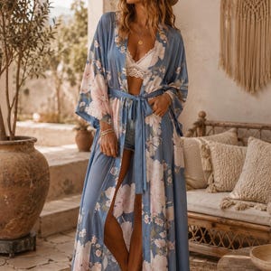 Peut inclure: Un peignoir long de style duster, ouvert sur le devant, dans une teinte bleu poussiéreux, orné d'un grand imprimé floral rose pâle et blanc. Le vêtement présente des manches larges et fluides, une ceinture assortie et deux poches latérales.