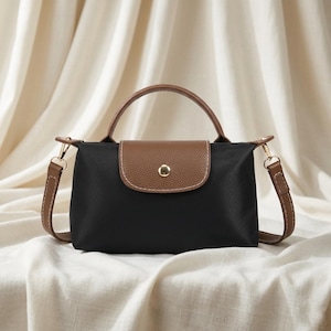 May include: Black handbag with a brown flap and handle. The bag has a gold-colored clasp and a detachable brown shoulder strap. The bag is made of a nylon-like material.