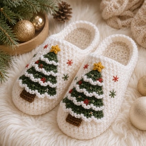 Puede incluir: Un par de pantuflas de ganchillo color blanco roto con una aplicación de árbol de Navidad verde oscuro en cada punta. Los árboles están rematados con una pequeña estrella amarilla y decorados con diminutos copos de nieve bordados.