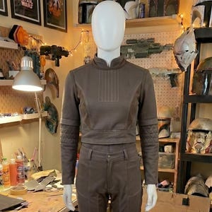 May include: A brown two-piece outfit consisting of a long-sleeved cropped top with a high collar and quilted elbow patches, paired with matching trousers featuring a button closure, belt loops, and reinforced knee panels with visible stitching.