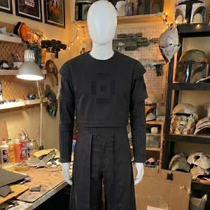 May include: A black costume set on a mannequin featuring a short-sleeved top with a square chest emblem, long-sleeved pinstriped undershirt, and matching trousers with an attached front skirt panel. The ensemble is entirely dark in color.