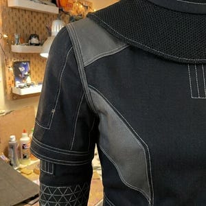 Puede incluir: Una prenda negra de manga larga con cuello alto y costuras blancas, un panel de hombro de malla texturizada y detalles en tela gris. La manga incluye un patr&oacute;n geom&eacute;trico triangular cerca del pu&ntilde;o y costuras reforzadas.