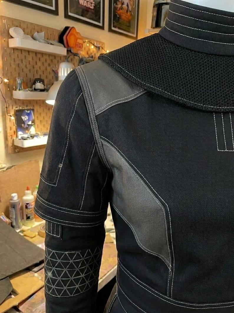 Puede incluir: Una prenda negra de manga larga con cuello alto y costuras blancas, un panel de hombro de malla texturizada y detalles en tela gris. La manga incluye un patr&oacute;n geom&eacute;trico triangular cerca del pu&ntilde;o y costuras reforzadas.