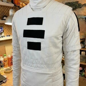 Puede incluir: Una t&uacute;nica blanca de manga larga con cuello alto y tres parches rectangulares negros horizontales centrados en el pecho. La prenda presenta costuras horizontales acolchadas en el torso y las mangas, con un dise&ntilde;o estructurado.