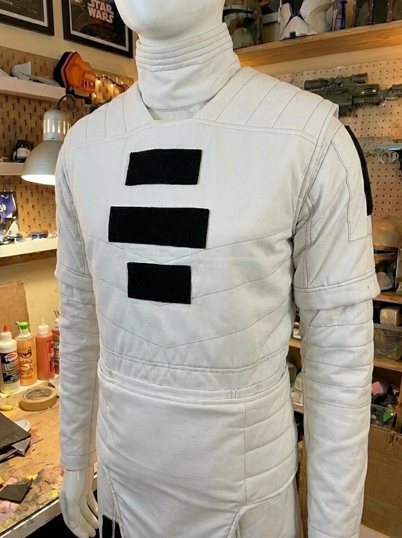 Puede incluir: Una t&uacute;nica blanca de manga larga con cuello alto y tres parches rectangulares negros horizontales centrados en el pecho. La prenda presenta costuras horizontales acolchadas en el torso y las mangas, con un dise&ntilde;o estructurado.