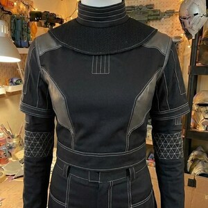 Puede incluir: Una prenda superior negra de manga larga con cuello alto, que presenta costuras blancas en contraste, un panel texturizado en el canes&uacute; y patrones geom&eacute;tricos triangulares en los antebrazos. La prenda incluye detalles estructurados en los hombros.