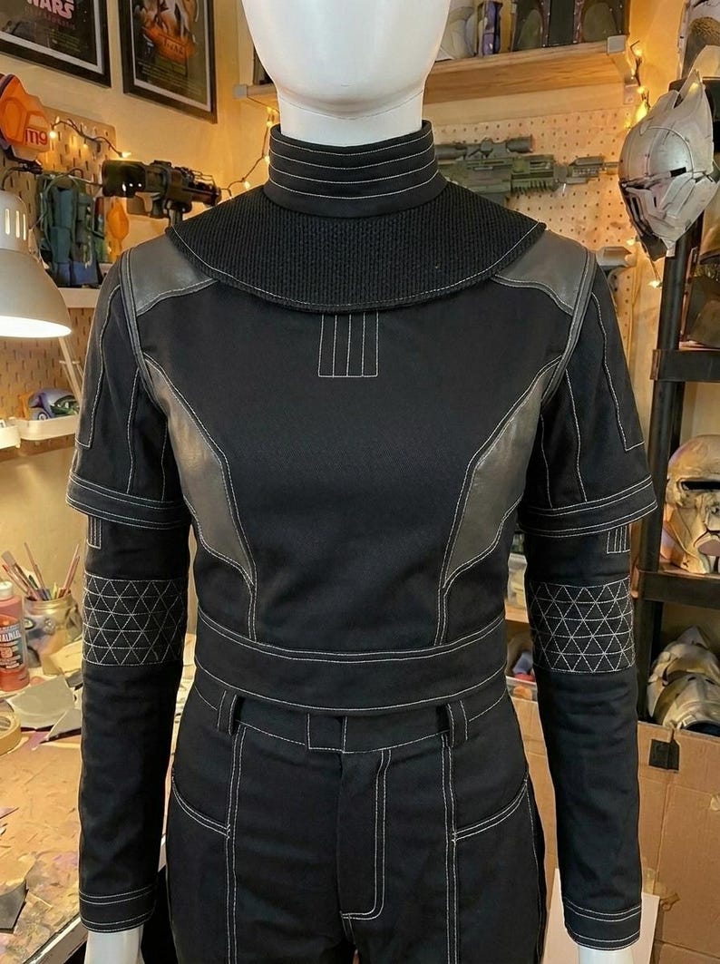 Puede incluir: Una prenda superior negra de manga larga con cuello alto, que presenta costuras blancas en contraste, un panel texturizado en el canes&uacute; y patrones geom&eacute;tricos triangulares en los antebrazos. La prenda incluye detalles estructurados en los hombros.