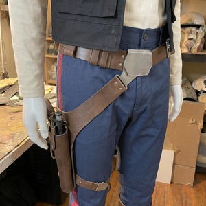 May include: A costume ensemble featuring a cream long-sleeve shirt, a black vest, and navy blue trousers with red side stripes. A brown leather utility belt with a metallic buckle and a matching drop-leg holster containing a black blaster pistol.