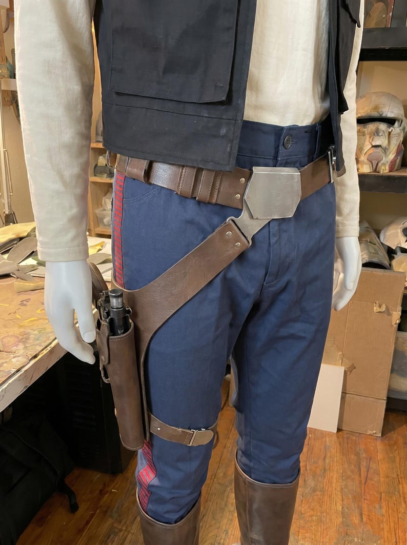 May include: A costume ensemble featuring a cream long-sleeve shirt, a black vest, and navy blue trousers with red side stripes. A brown leather utility belt with a metallic buckle and a matching drop-leg holster containing a black blaster pistol.