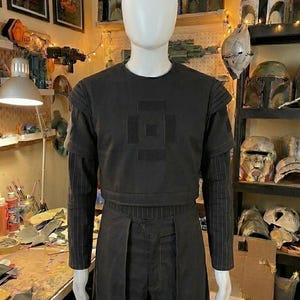 May include: A black short-sleeved tactical vest with a square patch design on the chest, worn over a long-sleeved shirt with thin vertical pinstripes. The outfit includes matching black trousers with a structured, layered front panel design.