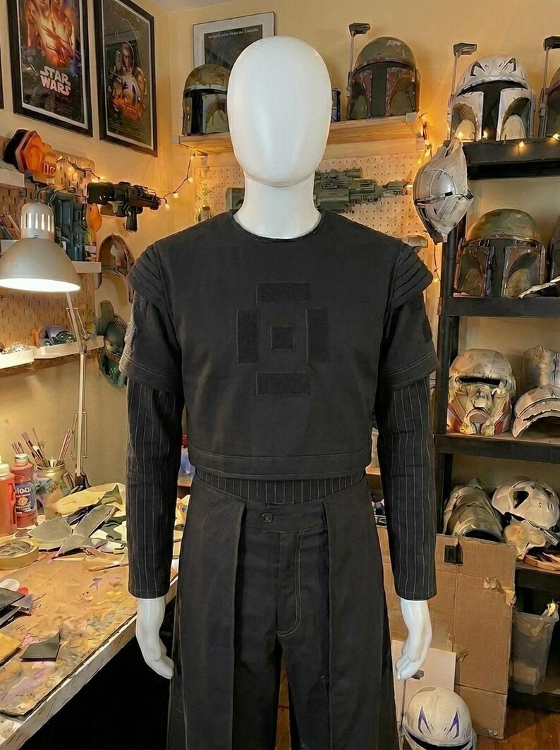 May include: A black short-sleeved tactical vest with a square patch design on the chest, worn over a long-sleeved shirt with thin vertical pinstripes. The outfit includes matching black trousers with a structured, layered front panel design.