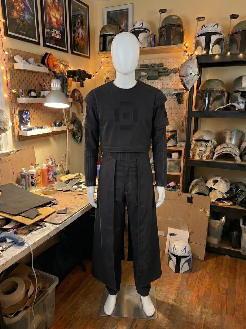 May include: A black costume set on a mannequin featuring a short-sleeved top with a square chest emblem, long-sleeved pinstriped undershirt, and matching trousers with an attached front skirt panel. The ensemble is entirely dark in color.