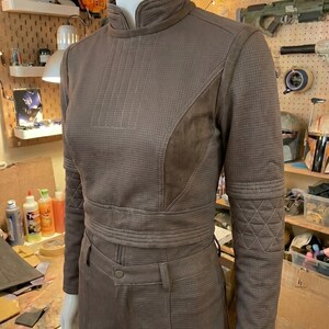May include: A brown long-sleeved jacket and matching pants set featuring a textured, perforated fabric finish. The jacket has a high collar, a front zipper, and quilted diamond-patterned detailing on the forearms, worn on a mannequin.