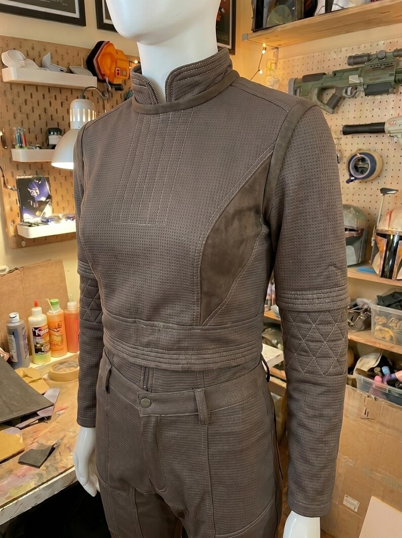 May include: A brown long-sleeved jacket and matching pants set featuring a textured, perforated fabric finish. The jacket has a high collar, a front zipper, and quilted diamond-patterned detailing on the forearms, worn on a mannequin.