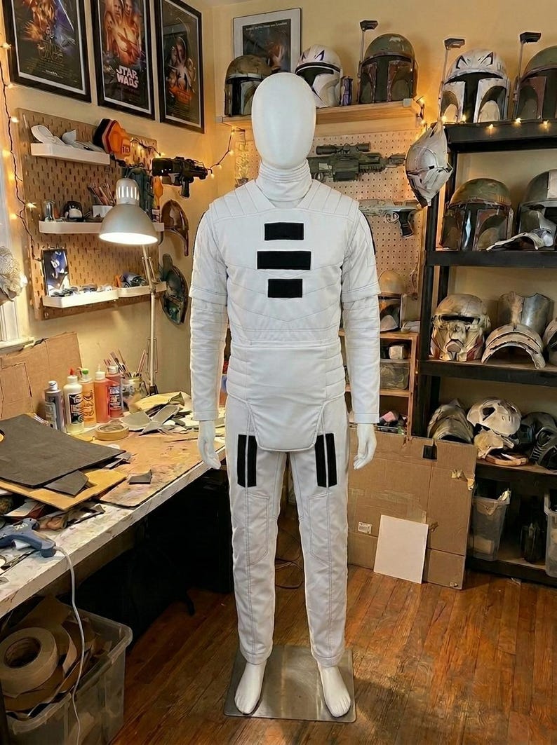 Puede incluir: Un traje de cuerpo completo de color blanco con cuello alto, textura acolchada en el pecho y los hombros, y un panel liso en la cintura. La prenda incluye tres parches rectangulares negros horizontales en el pecho y dos rayas negras verticales en cada pierna.