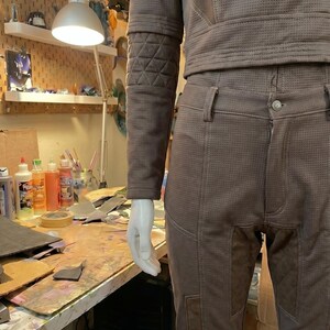 May include: A taupe-colored long-sleeve top and matching pants set featuring a micro-perforated texture throughout. The garment includes quilted panel detailing on the sleeves and thighs, visible white stitching, and a button closure on the pants.