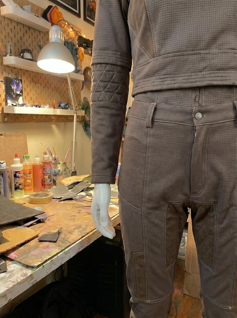May include: A taupe-colored long-sleeve top and matching pants set featuring a micro-perforated texture throughout. The garment includes quilted panel detailing on the sleeves and thighs, visible white stitching, and a button closure on the pants.