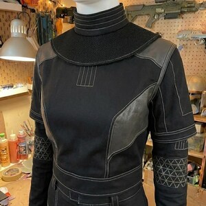 Puede incluir: Una camisa t&aacute;ctica negra de manga larga con cuello alto, un canes&uacute; de malla texturizada y paneles grises de aspecto cuero en el pecho y los hombros. Las mangas incluyen secciones con patrones geom&eacute;tricos y costuras decorativas blancas.