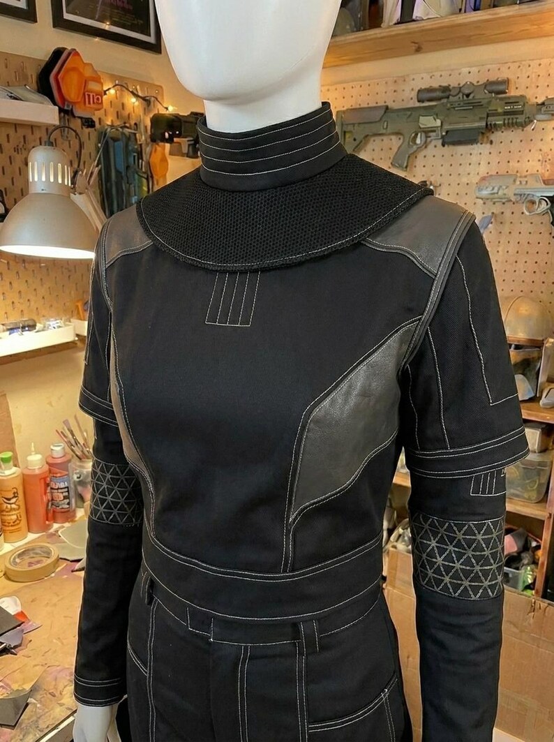 Puede incluir: Una camisa t&aacute;ctica negra de manga larga con cuello alto, un canes&uacute; de malla texturizada y paneles grises de aspecto cuero en el pecho y los hombros. Las mangas incluyen secciones con patrones geom&eacute;tricos y costuras decorativas blancas.