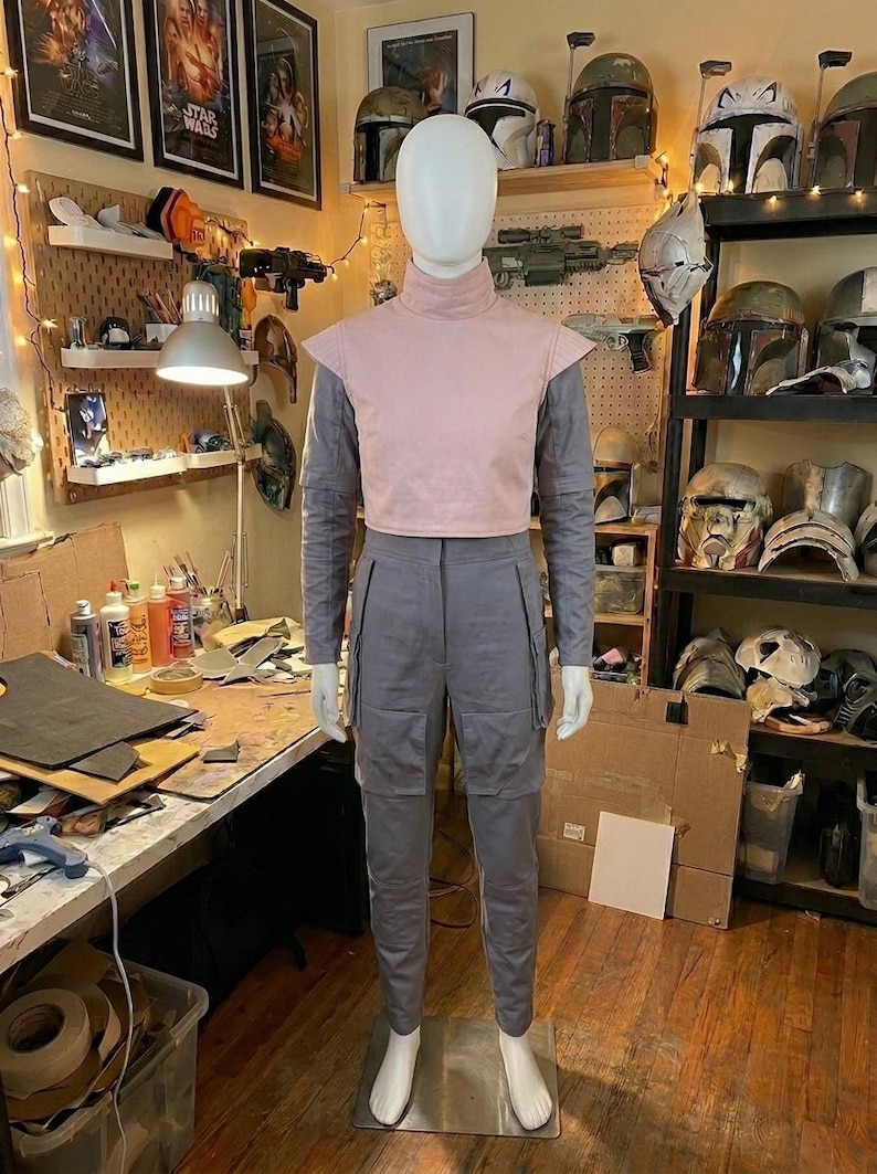 K&ouml;nnte beinhalten: Eine Schaufensterpuppe tr&auml;gt ein hellrosa &auml;rmelloses Oberteil mit Stehkragen &uuml;ber einem grauen Langarmshirt und einer passenden grauen Cargohose mit gro&szlig;en Seitentaschen. Das Outfit zeichnet sich durch ein strukturiertes Design mit klaren Linien aus.