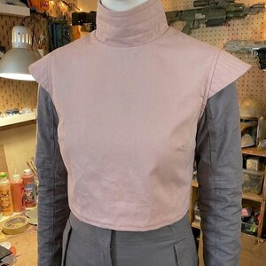 K&ouml;nnte beinhalten: Eine hellrosa &auml;rmellose Weste mit hohem Kragen und strukturierten Schulterpartien, getragen &uuml;ber einem grauen Langarmshirt und einer grauen Hose. Die Weste weist horizontale N&auml;hte am Kragen und an den Schultern auf, was einen Lagen-Look erzeugt.