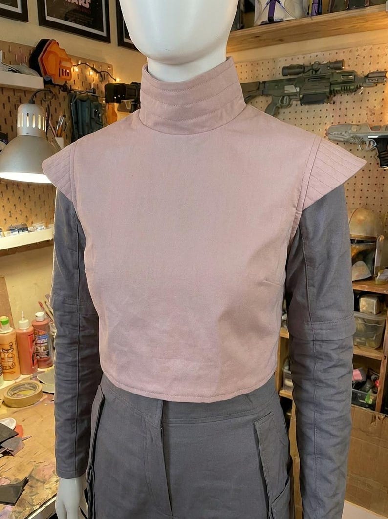 K&ouml;nnte beinhalten: Eine hellrosa &auml;rmellose Weste mit hohem Kragen und strukturierten Schulterpartien, getragen &uuml;ber einem grauen Langarmshirt und einer grauen Hose. Die Weste weist horizontale N&auml;hte am Kragen und an den Schultern auf, was einen Lagen-Look erzeugt.