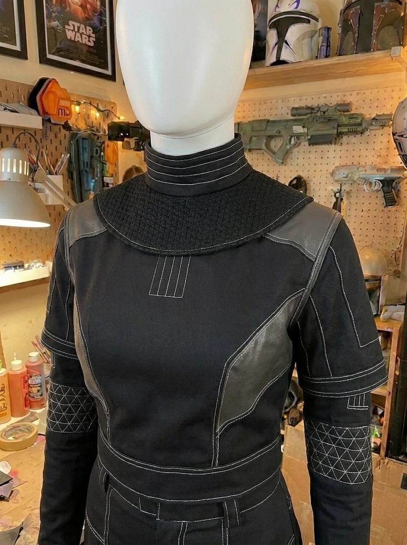 Puede incluir: Una prenda superior negra de manga larga con cuello alto, que presenta un canes&uacute; negro texturizado, paneles grises en los hombros y costuras blancas en contraste. Las mangas incluyen patrones geom&eacute;tricos acolchados en los antebrazos.
