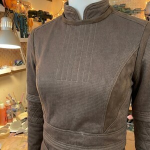 May include: A brown long-sleeved top with a high collar, featuring a textured micro-perforated fabric, vertical stitched detailing on the chest, and quilted diamond patterns on the upper sleeves, worn on a white mannequin torso.